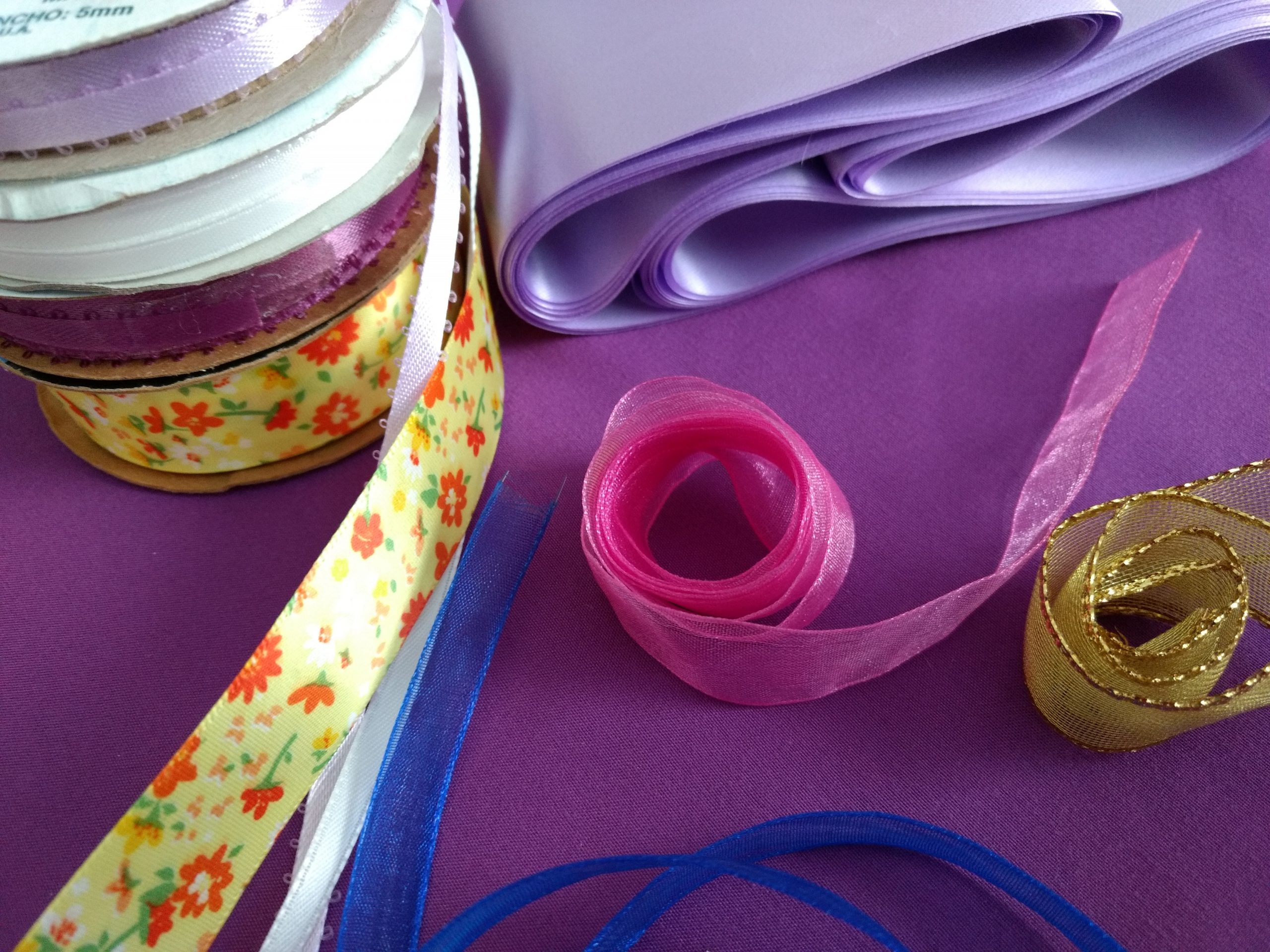 A white, purple, blue, gold, pink, and flowery ribbon roll along with a purple sheet, set against a purple background